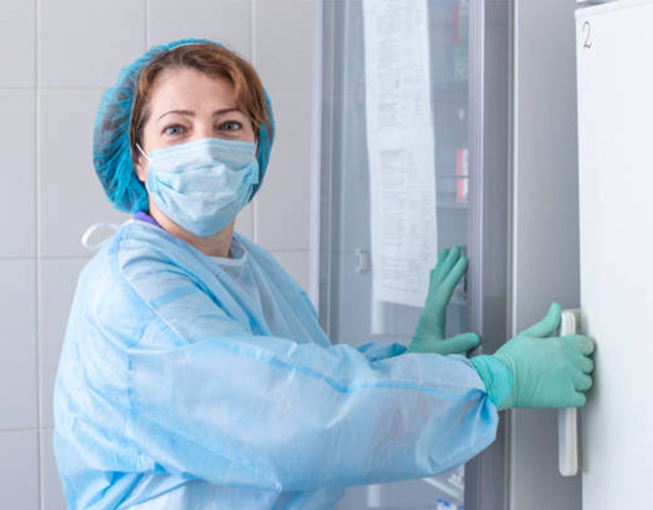 nurse in a medical cap, mask and blue protective gown opens the refrigerator door with the vaccine. Cabinet for procedures and vaccinations. Women work in healthcare