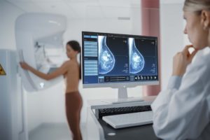 Breast Cancer Computer Screen in Hospital Radiology Room: Beautiful Multiethnic Adult Woman Standing Topless Undergoing Mammography Screening Procedure. Screen Showing the Mammogram Scans of Dense Breast Tissues.